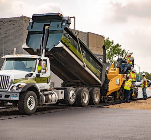 Truck loading paver with hot asphalt mix at a commercial parking lot project.