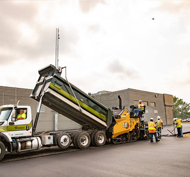 Tandem truck loading hot asphalt into paver on a commercial paving site.