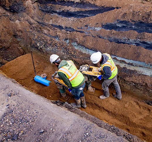 Two men compacting soil overtop underground utilities with rammer compactors.