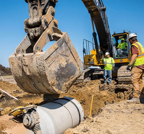 Excavator bucket unloading dirt on underground utility pipe.