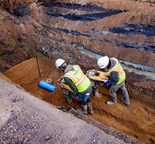 Two men compacting soil overtop underground utilities with rammer compactors.