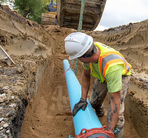 A construction worker in a hard hat and safety vest installs a large blue pipe in a deep, sandy trench at an outdoor worksite, with an excavator in the background.