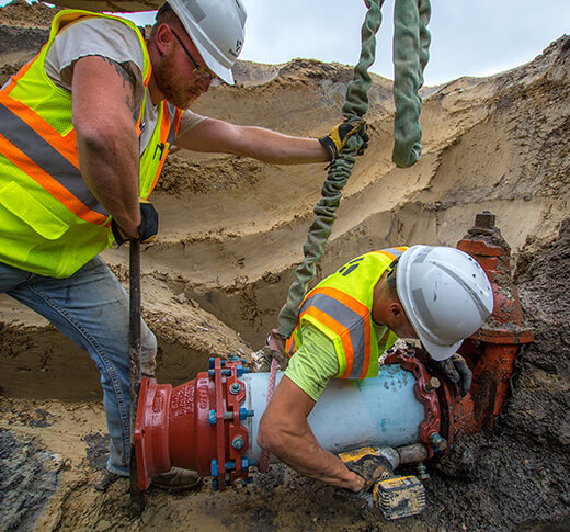 Two construction workers in safety vests and helmets are installing or repairing a large pipeline in a sandy trench, using tools and lifting straps to position the pipe and attach fittings.