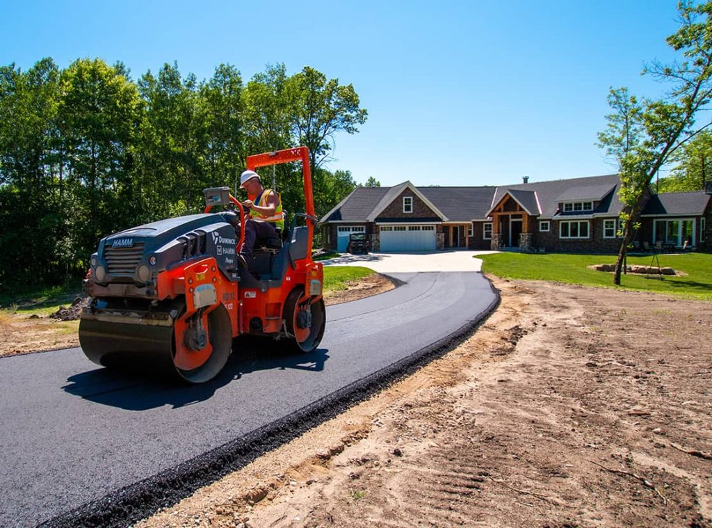 A construction worker drives a steamroller over fresh asphalt, paving a curving driveway leading to a large house surrounded by trees on a sunny day.