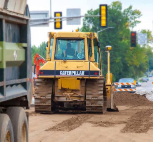 A yellow Caterpillar bulldozer spreads dirt at a road construction site near traffic lights, with road barriers and trees in the background and a truck partially visible on the left.