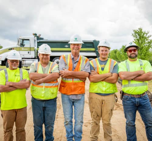 Safety gear and hard hats stand with arms crossed on a work site, smiling at the camera. A large truck and green trees are visible in the background under a cloudy sky.