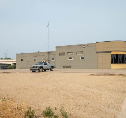 A pickup truck is parked on a gravel lot in front of a large, flat-roofed beige and gray building with glass windows on one corner under a clear sky. A few small buildings are visible in the background.