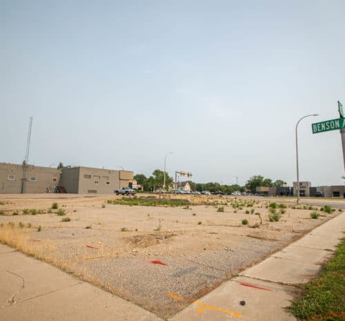 A vacant, cracked parking lot with weeds growing through the pavement sits at the corner of Benson Ave S.W., next to a gray building and a red fire hydrant under a hazy sky.