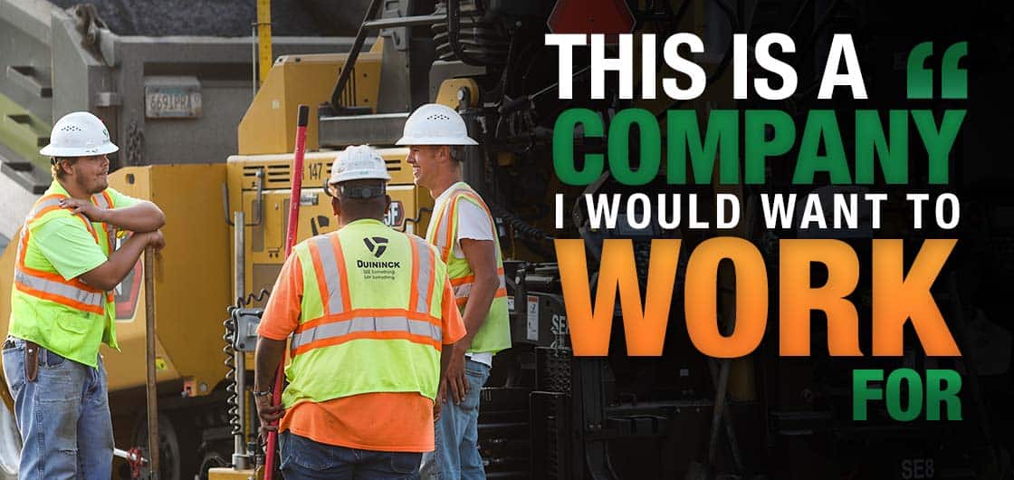 Three men in safety vests and hard hats in front of a paver talking.