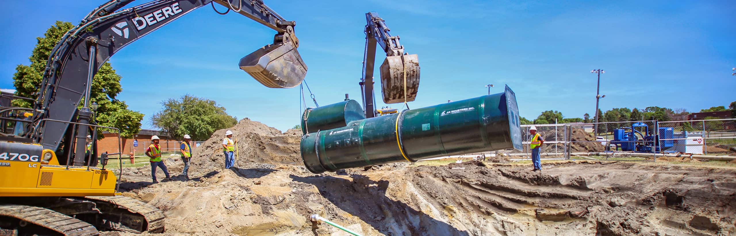 Two John Deer excavators lift a large sanitary lift station over a trench.