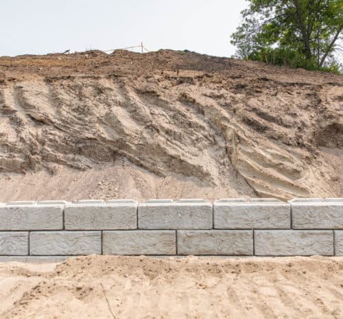 A retaining wall made of large concrete blocks stands in front of a steep, eroded dirt hillside with sparse vegetation at the top.