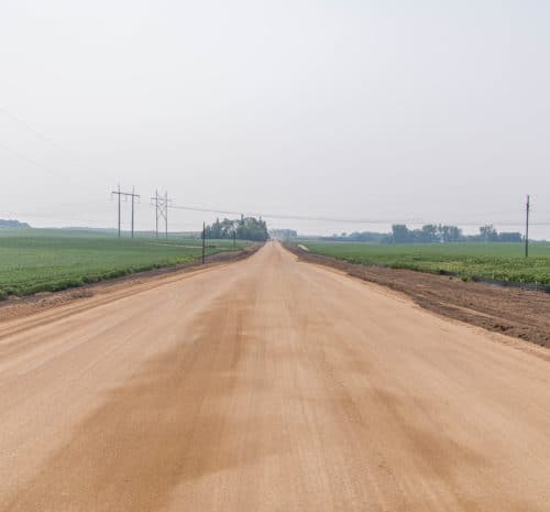 A straight, wide dirt road runs through flat farmland with green crops on both sides, under a hazy sky. Power lines parallel the road. Trees are visible in the distance.