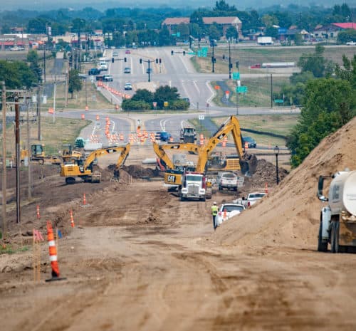 A construction site with several excavators, trucks, and workers on a dirt road near a busy intersection. Traffic cones, dirt piles, and construction equipment are visible, with buildings and trees in the background.