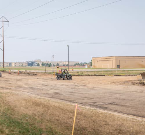 Construction site with dirt ground, construction vehicles, and large pipes. Buildings and power lines are visible in the background under a hazy sky. Pink stakes mark the area in the foreground.