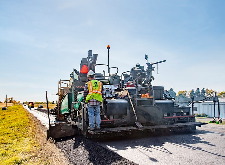 Screed operator standing on paver while paving a government highway.
