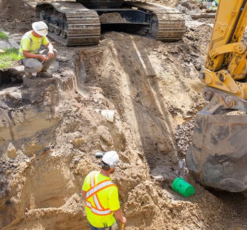 One man kneeling overtop a trench talking to another man in the trench of an underground construction site.