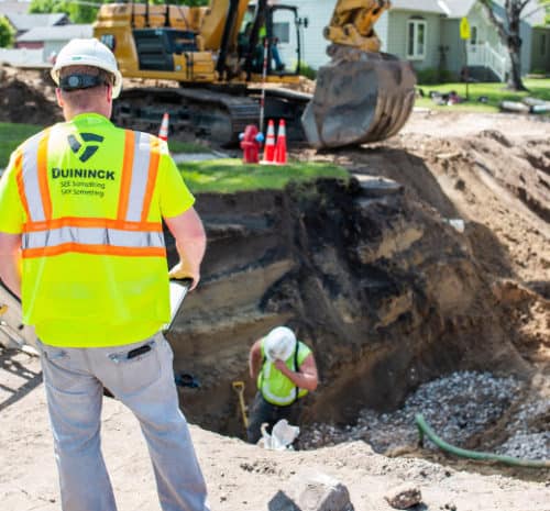 A man in a safety vest observes from overtop of a trench of an underground construction site.