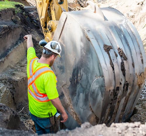 Man in safety vest and hard hat directing a large excavator in trench.