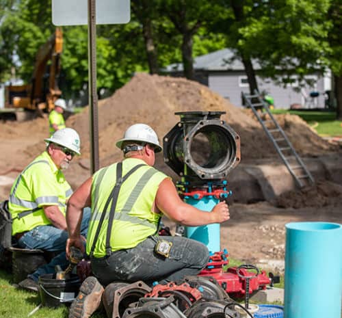 Two men in safety gear and hard hats connect a water main near an underground construction site.