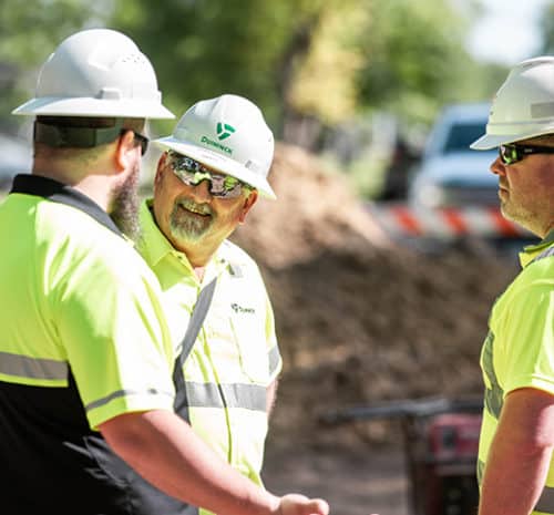 Three men in safety vests and hard hats talking at a construction site.