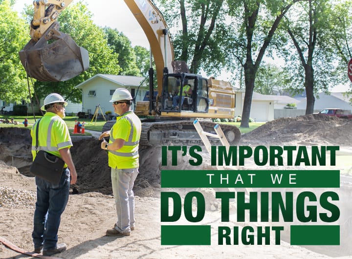 Two men in high-visibility safety gear talking at a construction site with an excavator in the background.