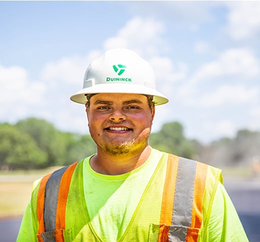 A construction worker wearing a white hard hat and a neon yellow safety vest with orange stripes smiles outdoors on a sunny day, with trees and a blue sky in the background.