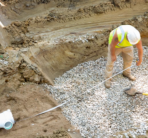 Man in helmet measures a trench with a tape measure beside a gravel bed and a drainage pipe. The trench shows layers of soil. - July 10, 2020