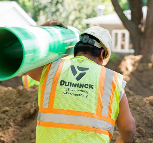A construction worker in a reflective vest and hard hat carries a large green pipe on their shoulder at an outdoor worksite. The vest reads DUININCK. SEE Something SAY Something.