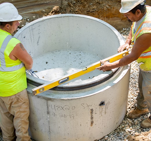 Two construction workers in safety gear use a level tool to check the alignment of a large concrete cylinder at a construction site with gravel on the ground.