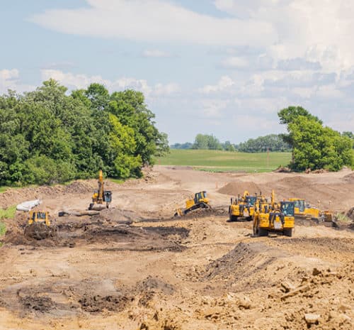 Excavators work on a large, dirt-filled construction site, showcasing the busy construction activity amid green trees and fields under a partly cloudy sky.