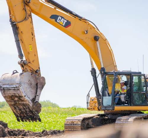 A construction worker operates a large yellow Caterpillar excavator, digging into soil on a sunny day with green fields in the background, showcasing the essential role of construction in shaping our environment.