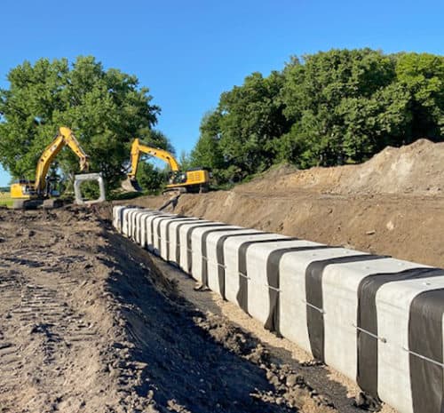 Two yellow excavators work at a construction site, installing large rectangular concrete blocks in a trench amid dirt piles and green trees, showcasing the busy construction scene under a clear blue sky.