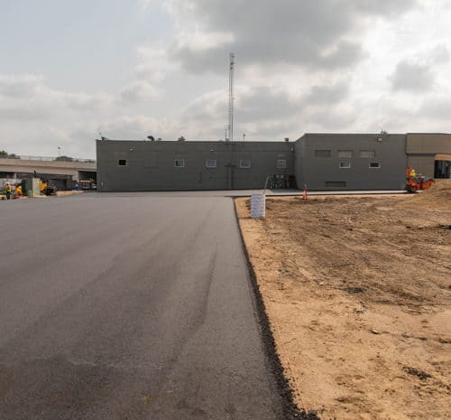Freshly paved asphalt road next to a construction site with exposed soil, leading towards a gray industrial building. Construction workers and equipment are visible near the building under a cloudy sky.