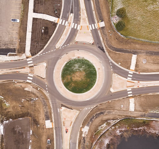 Aerial view of a roundabout with four roads connecting to it, crosswalks at each entrance, and a grassy center. Surrounding areas include grass, parking lots, and some construction.