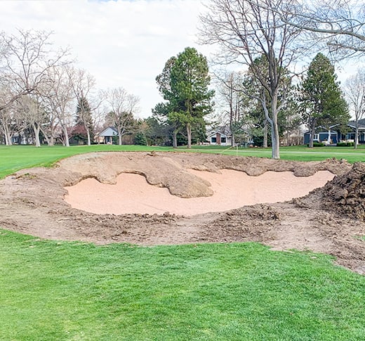 A golf course bunker under renovation, with exposed dirt edges and fresh sand, surrounded by green grass, leafless trees, and houses in the background.