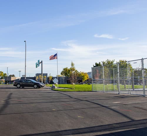 A mostly empty parking lot with a single black car, an American flag on a pole, a chain-link fence, and some green trees under a clear blue sky.