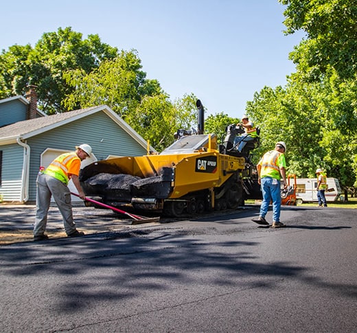 Four construction workers in safety vests and hats are paving a residential street with a yellow asphalt paver, surrounded by trees and houses on a sunny day.