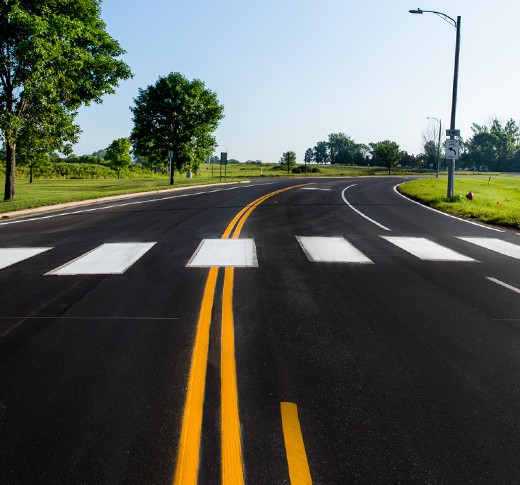 A freshly paved road with bright yellow lane markings and a white pedestrian crosswalk curves to the right through a green, tree-lined area under a clear blue sky.