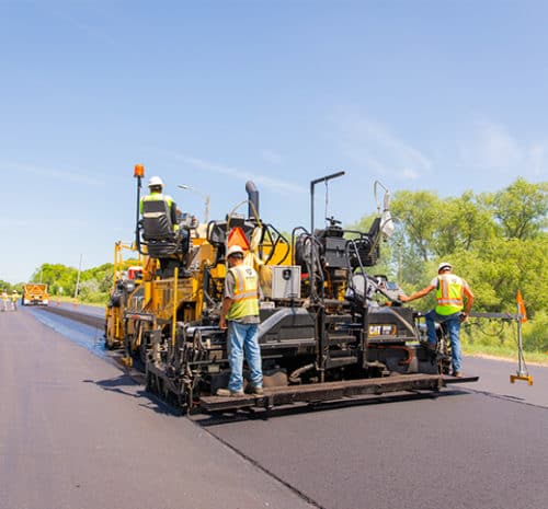 asphalt paving machine on a sunny day resurfacing a road surrounded by green trees and grass.