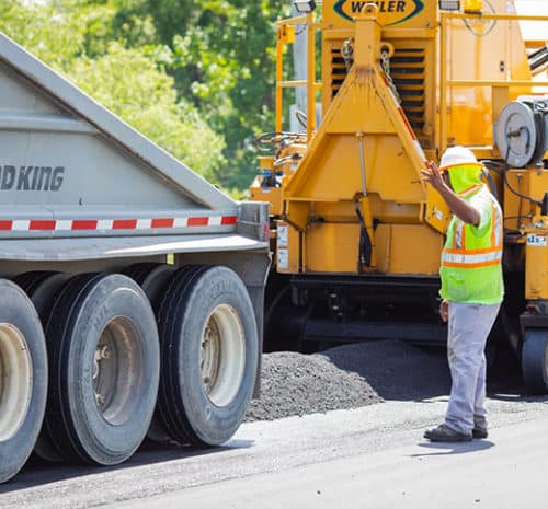 Construction worker wearing a safety vest and helmet signals to a dump truck driver next to a road paving machine, with fresh asphalt being laid on a sunny day.