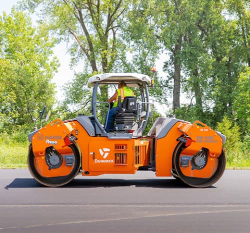 A worker operates an orange road roller on freshly paved asphalt with green trees and foliage in the background.