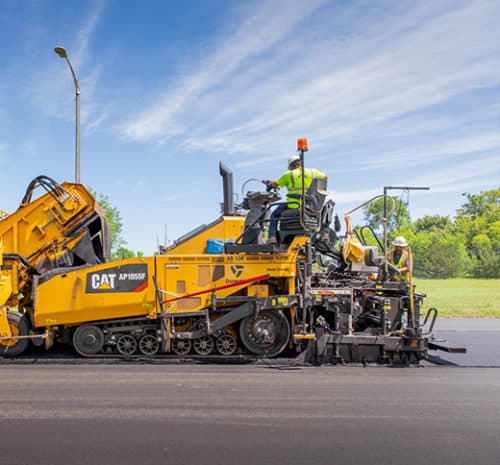Two workers operate a large yellow Caterpillar asphalt paving machine on a road under a blue sky, with grass and trees in the background.