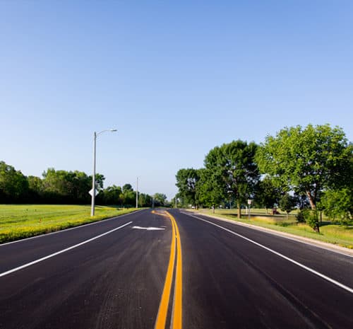 A freshly paved two-lane road with bright yellow center lines curves gently, bordered by green grass and trees under a clear blue sky.