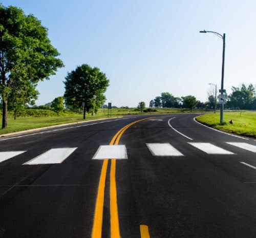 A freshly paved road with a yellow double line curves right, passing a crosswalk. Green grass, trees, and a clear blue sky border the street.