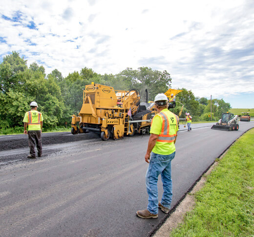Road construction crew wearing safety vests and helmets operate heavy machinery to lay fresh asphalt on a rural road bordered by green trees under a partly cloudy sky.