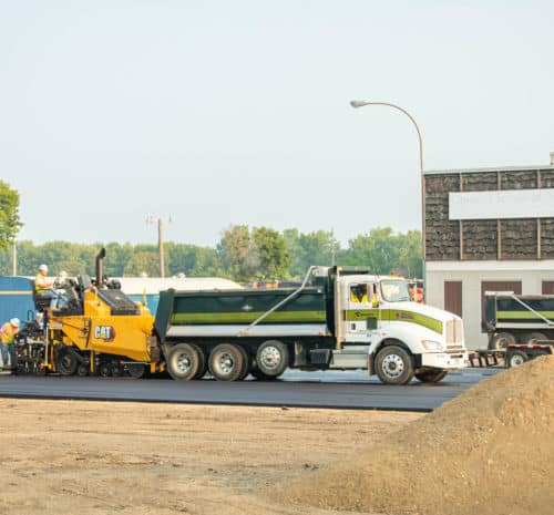 Construction workers in safety vests operate a yellow asphalt paving machine next to a large green and white dump truck on a street, with a dirt pile in the foreground and buildings and trees in the background.