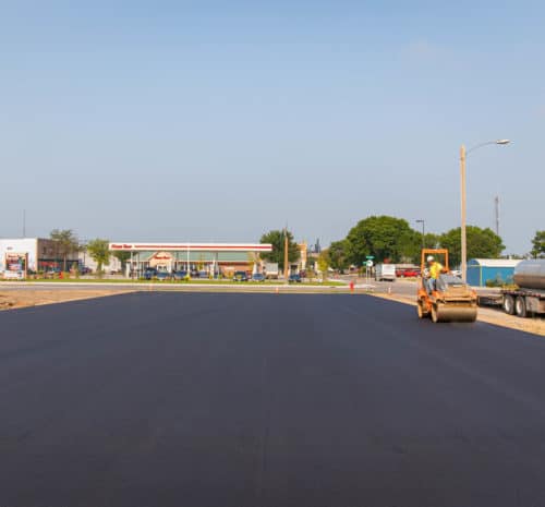 A freshly paved road with a construction worker operating a steamroller on the right, a white truck parked nearby, and a gas station and trees visible in the background under a clear sky.