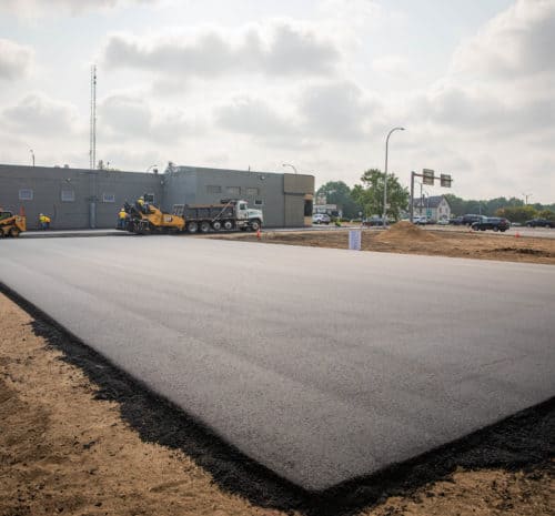 Freshly paved asphalt parking lot with construction equipment and workers near a gray building; the surrounding area has exposed dirt, and traffic is visible in the background under a cloudy sky.