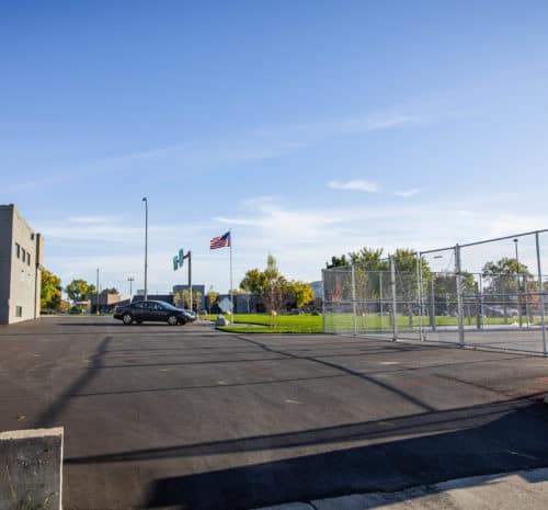 A parking lot with several parked vans and a black car, bordered by a chain-link fence. An American flag flies in the background - 2021