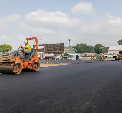 A worker operates an orange and black road roller to smooth freshly paved asphalt in a parking lot under a partly cloudy sky, with other workers and vehicles in the background.